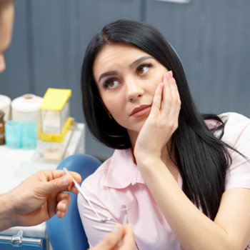 black haired woman holding her cheek sitting in a dental chair