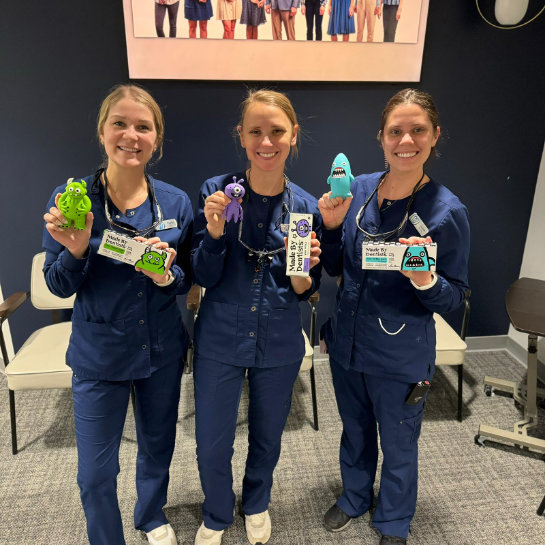 Three nurses in blue scrubs holding up toys in an office.